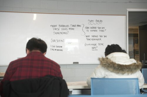 The image depicts a classroom setting with two individuals seated at a table, facing a whiteboard. The individuals are seen from behind; one is wearing a red plaid shirt, and the other is in a white jacket with a fur-lined hood. The focus is on the whiteboard, which contains handwritten text in black marker.

The text on the whiteboard includes several sections. One section titled 'COPE AHEAD' suggests strategies or questions like 'How would you do to handle this?' and 'What would you say was your experience?' Another section discusses 'Goal Setting,' listing 'Long term' and 'Like a mountain.' The phrase 'What barriers could make these goals more challenging?' is also visible.

The room has a modern, educational atmosphere, with light-colored walls and a bright, evenly lit environment. The scene suggests an educational or workshop context, possibly focusing on personal development or therapeutic strategies. The setting is calm and conducive to learning or discussion, with a focus on planning and coping strategies.