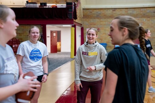 The image depicts a group of young women in a gymnasium setting, engaging in a casual conversation or possibly a sports-related activity. The gym has a polished floor with maroon and beige tones, and a brick wall in the background, indicating an indoor sports facility. 

One woman, wearing a light gray hoodie with 'ROLL COBBS' printed in maroon and gold, stands out as she smiles and interacts with others. This text suggests a connection to a sports team or school spirit, possibly related to a college or high school team. Another woman holds a football, indicating the activity might be related to a football practice or training session.

The individuals are dressed in casual athletic wear, suggesting a relaxed, informal atmosphere. The lighting is bright and natural, contributing to a lively and energetic environment. The overall mood is friendly and collegial, with the participants appearing engaged and comfortable.

This setting and attire suggest a practice or team-building event, possibly linked to a school or community sports program. The presence of sports equipment and the gymnasium context emphasize a focus on athletic activities.