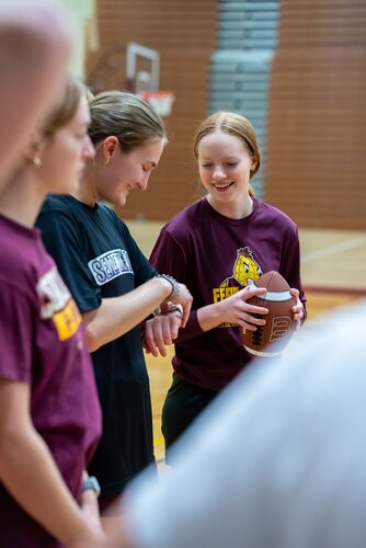 The image depicts a group of young individuals in a gymnasium setting, engaged in a casual or practice session. The focus is on two people in the foreground. One person, wearing a maroon shirt with a yellow logo, is holding an American football and smiling, suggesting a friendly or instructional interaction. Another person, in a black shirt, is looking at their wristwatch, possibly indicating a timing or scheduling aspect of the activity.

The gymnasium features wooden flooring and a basketball hoop in the background, indicating a multi-sport facility. The lighting is bright and even, typical of indoor sports venues. The atmosphere appears relaxed and collegial, with participants seemingly enjoying the activity.

The maroon shirts with logos suggest a team or school affiliation, possibly related to a sports team or physical education class. The presence of the football indicates a focus on American football, and the casual attire of the individuals suggests a practice or informal gathering rather than a competitive event.