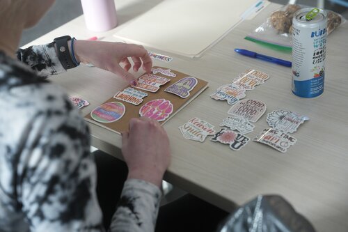 The image depicts a person engaged in a creative activity, arranging various colorful stickers on a flat surface, possibly a piece of cardboard or a folder. The stickers feature a variety of motivational and positive phrases in vibrant colors, such as 'Kindness is Magic,' 'Stay Positive,' and 'You Got This.' The person is wearing a black and white tie-dye long-sleeve shirt and a blue wristband.

On the table, there is a blue pen, a can of drink labeled 'Vita Coco,' and a partially visible plastic bag containing what appears to be cookies or snacks. The setting suggests a casual, relaxed environment, possibly a workshop or a personal crafting session.

The lighting is soft and natural, contributing to a calm and focused atmosphere. The activity conveys a sense of creativity and positivity, emphasizing themes of self-expression and encouragement. The overall scene suggests a personal or small group setting, where individuals are engaged in crafting or organizing inspirational materials.