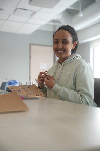 The image features a person sitting at a table in a modern, well-lit room. The individual is wearing a light-colored hoodie and has a cheerful expression, suggesting a relaxed and positive atmosphere. They are engaged in a creative activity, holding a small object, possibly a crafting tool or piece, while working on a project involving a notebook or scrapbook on the table. The notebook is adorned with various stickers or decorations, indicating a personal or artistic endeavor.

The setting appears to be a casual, indoor environment, possibly a classroom, workshop, or community center, as suggested by the presence of other crafting materials and a bright, airy space with large windows. The predominant colors in the image are soft and neutral, with the light gray walls and ceiling contributing to a calm and inviting ambiance.

The overall theme of the image conveys creativity, leisure, and personal expression, capturing a moment of focused yet enjoyable activity. The presence of crafting supplies and the individual's engagement with the project suggest a context of learning or artistic exploration, typical of a workshop or educational setting.