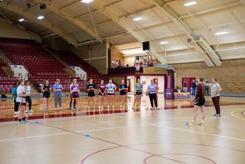 The image depicts a group of individuals in a gymnasium setting, likely participating in a sports or physical education activity. The gym features a high, arched ceiling with exposed beams and a combination of brick and panel walls. The flooring is typical of a basketball court, marked with lines and circles, and there are several blue cones placed on the floor, possibly for drills or exercises.

In the background, there are rows of maroon bleachers, indicating this is a space designed for spectators during events. A basketball hoop is visible, suggesting the primary use of the gym is for basketball, although it may accommodate other sports.

The group consists of both men and women, casually dressed in athletic wear. They are standing in a line, facing a person who appears to be instructing or leading the activity. This individual is positioned slightly apart from the group, gesturing as if explaining or demonstrating something.

The lighting is bright and evenly distributed, typical of indoor sports facilities, providing a clear view of the entire space. The atmosphere seems focused and educational, with participants attentive to the instructions being given.

This setting is reminiscent of a school or community sports program, where participants are engaged in learning or practicing a sport. The image captures a moment of instruction, emphasizing teamwork, learning, and physical activity.