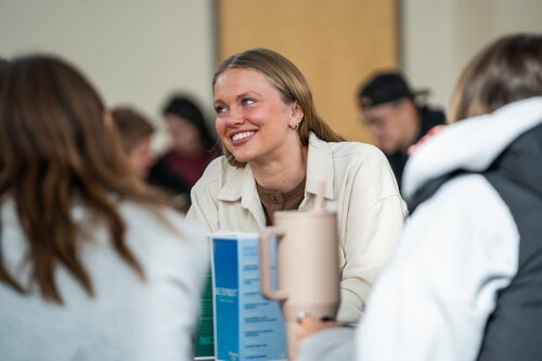 The image depicts a social setting, likely a casual gathering or meeting, where a group of people are seated around a table. The central focus is on a woman with light hair, wearing a white jacket, who is smiling and engaging with others. Her expression suggests a friendly and relaxed atmosphere. She is holding a beige mug, indicating a casual or informal setting, possibly a café or a community space.

The background shows other individuals, slightly out of focus, suggesting a busy environment with multiple conversations occurring simultaneously. The lighting is bright and natural, contributing to the warm and inviting ambiance. The presence of a menu or informational booklet on the table hints at a dining or communal event.

The setting and attire of the individuals suggest a contemporary, everyday context, with no explicit cultural or historical references. The overall mood is positive and sociable, highlighting interaction and connection among the participants.
