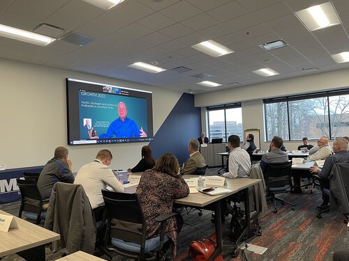 The image depicts a professional meeting or conference setting within a modern, well-lit room. Attendees are seated around several rectangular tables arranged in a U-shape, facing a large screen at the front of the room. The screen displays a virtual presentation featuring a speaker in a blue shirt, with the text "GROWTH 2022" visible. The speaker appears to be addressing the audience, suggesting a focus on business or professional development.

The room features a neutral color palette with white walls, a gray carpet with red accents, and large windows allowing natural light to enter. The ceiling has recessed lighting, contributing to the bright atmosphere. Participants are dressed in business attire, indicating a formal or corporate environment.

The overall mood is professional and focused, with attendees engaged in the presentation. The setting suggests a seminar or workshop, possibly related to business growth strategies or leadership training. The presence of both in-person and virtual elements highlights a hybrid meeting format, common in contemporary professional environments.