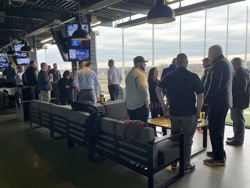 The image depicts a social gathering at a modern indoor golf venue, likely a driving range with an entertainment focus. The setting features large floor-to-ceiling windows, allowing natural light to illuminate the space and providing a view of the outdoor golf area. The interior is designed with sleek, contemporary furniture, including cushioned sofas and high tables, creating a casual and relaxed atmosphere.

Several groups of people are engaged in conversation, suggesting a networking or social event. The individuals are dressed in a mix of casual and business-casual attire, indicating a blend of leisure and professional interaction. Some people are holding drinks, enhancing the social aspect of the gathering.

Multiple flat-screen TVs are mounted on the walls, displaying sports content, adding to the entertainment environment. The lighting is a mix of natural daylight and artificial overhead lights, contributing to a bright and inviting ambiance.

The overall mood is lively and sociable, with a focus on leisure and interaction. The venue is reminiscent of popular entertainment golf destinations like Topgolf, known for combining sports with social activities. The setting suggests a space designed for both recreation and informal business networking, making it a versatile location for various events.