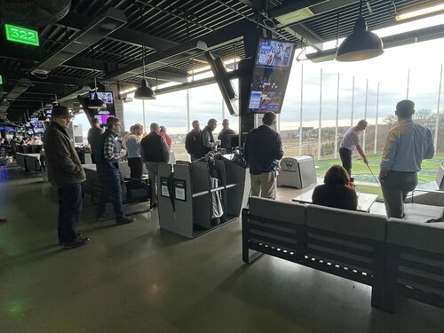 The image depicts an indoor golf entertainment venue, likely a Topgolf location, characterized by its distinctive driving range setup. Several individuals are gathered around a series of golf bays, each equipped with seating and tables. One person is actively swinging a golf club, aiming towards the outdoor range visible through large, netted openings. The scene is bustling with activity, as groups of people are engaged in conversation or observing the golfer.

The setting features modern, industrial-style architecture with exposed beams and overhead lighting. Multiple flat-screen TVs are mounted throughout the venue, displaying various content, enhancing the sports entertainment atmosphere. The lighting is a mix of natural daylight streaming in from outside and artificial illumination from the overhead fixtures.

The predominant colors include neutral tones from the interior, contrasted by the green of the outdoor turf. The atmosphere is lively and social, suggesting a recreational outing or corporate event. The digital clock displays the time as 3:22, adding a sense of real-time engagement to the scene. The image captures a blend of leisure and social interaction, typical of a modern entertainment complex designed for group activities.