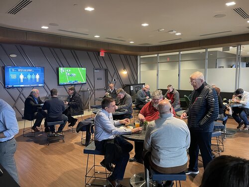 The image depicts a modern, indoor lounge or sports bar setting with a group of people engaged in conversation and leisure activities. The room features contemporary design elements, such as sleek wooden flooring and geometric wall patterns. Two large flat-screen televisions are mounted on the walls, displaying different content. One screen shows a sports game, likely American football, while the other displays informational graphics or statistics.

The individuals, predominantly middle-aged men, are casually dressed, with some wearing jackets or sweaters. They are seated at small, round tables, engaging in discussions or enjoying food and drinks. The atmosphere is relaxed and social, with a sense of camaraderie among the attendees.

The lighting is bright and evenly distributed, contributing to the open and inviting ambiance. The space appears to be part of a larger venue, possibly a conference center or a hospitality suite at a sports event, given the presence of the TVs and the casual, yet business-like attire of the guests.

Overall, the image conveys a theme of social interaction and leisure in a professional yet relaxed environment, possibly related to a sporting event or corporate gathering.