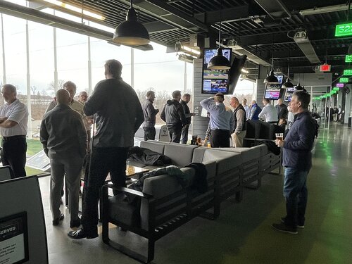 The image depicts a group of people gathered in an indoor golf facility, likely a driving range. The setting features a modern, industrial design with high ceilings and exposed beams. Large windows on the left side offer a view of the outdoor range, allowing natural light to illuminate the space. 

The individuals, mostly men, are dressed in casual to business-casual attire, suggesting a corporate or social event. They are engaged in conversation, some holding drinks, creating a relaxed and social atmosphere. A few people are focused on the golf activity, either preparing to swing or observing others.

The area is furnished with comfortable seating, including couches and tables, indicating a lounge-like environment. Multiple television screens are mounted above, displaying sports channels, adding to the recreational ambiance.

The lighting is a mix of natural daylight and artificial lighting from overhead fixtures, contributing to a bright and inviting setting. The overall mood is casual and sociable, with an emphasis on leisure and networking. The image captures a blend of sports and social interaction, typical of entertainment venues like Topgolf.