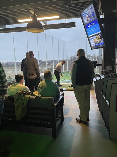 The image depicts an indoor golf entertainment venue, likely a Topgolf location, where a group of people is gathered. The setting is modern and casual, with a focus on recreational activity. 

In the foreground, several individuals are seated on a couch, watching someone actively engaged in swinging a golf club. This person is positioned at a tee, aiming towards an outdoor driving range visible through large, floor-to-ceiling windows. The outdoor area is enclosed with netting, typical of such venues to contain golf balls.

The lighting inside is dim, with overhead industrial-style pendant lights contributing to a relaxed atmosphere. The interior features a combination of concrete and metal, enhancing the contemporary design. 

On the right side of the image, a large digital screen displays various information, possibly related to the game or venue services, with blue and white as predominant colors. The screen also shows images of food and drinks, suggesting a social and leisure-focused environment.

The people in the image are dressed casually, indicating a laid-back, social gathering rather than a formal event. The overall mood is one of leisure and enjoyment, with participants likely engaging in friendly competition or simply enjoying the company and activity.

The setting and activity suggest a cultural context related to modern social entertainment, where sports and socializing are combined in a venue designed for both recreation and relaxation.