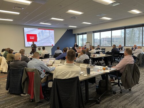 The image depicts a professional conference or seminar taking place in a modern, well-lit meeting room. The room is filled with attendees seated at several rectangular tables arranged in a U-shape, facing a large screen displaying a presentation slide titled "Current Reality." The setting suggests a corporate or educational environment, with participants engaged in listening to a speaker standing near the screen. 

The attendees, mostly dressed in business casual attire, are focused on the presentation, with some taking notes or looking at materials on the tables. The tables are equipped with nameplates, water bottles, and notepads, indicating an organized and formal setup. 

Large windows on one side of the room allow natural light to flood in, complementing the overhead fluorescent lighting, creating a bright and professional atmosphere. The view outside shows a glimpse of trees and a multi-story building, suggesting an urban or suburban location.

The overall mood is one of concentration and learning, typical of a business workshop or training session. The setting and arrangement convey a sense of collaboration and professional development.