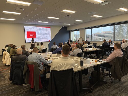 The image depicts a professional conference or seminar setting in a modern, well-lit room. The room is filled with attendees seated at tables arranged in rows, facing a large screen at the front. The screen displays a presentation slide with the title "Current Reality," indicating a focus on discussing present conditions or challenges. The room has a neutral color palette with gray carpeting and white walls, complemented by large windows on one side, allowing natural light to enter and providing a view of an office building and trees outside.

The attendees, predominantly dressed in business attire, are engaged in the presentation, with some taking notes or looking at materials on the tables. Each table has nameplates, suggesting a formal or organized event, possibly a workshop or corporate training session. The atmosphere is professional and focused, indicative of a business or educational environment.

The presenter stands near the screen, actively engaging with the audience, possibly explaining or elaborating on the slide content. The presence of water bottles, notebooks, and other personal items on the tables suggests a full-day event where participants are encouraged to stay hydrated and take notes.

Overall, the image conveys a theme of learning, collaboration, and professional development, set in a contemporary corporate environment.