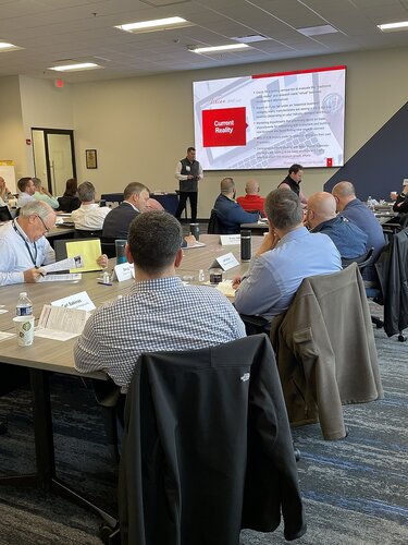 The image depicts a professional meeting or workshop taking place in a modern conference room. The setting includes a large group of individuals seated around several rectangular tables arranged in a U-shape. The participants are engaged in a presentation, focusing on a speaker standing at the front of the room. The speaker is positioned near a large digital screen displaying a slide titled "Current Reality," which includes text and a red square with white lettering.

The attendees are dressed in business casual attire, with some wearing jackets draped over their chairs. Papers, notebooks, and coffee cups are visible on the tables, indicating an interactive and attentive atmosphere. The room is well-lit with overhead lighting, and the walls are painted in neutral tones, contributing to a professional environment.

The carpet features a blue and gray pattern, adding a touch of color to the space. The overall mood is focused and collaborative, suggesting a setting where ideas are being exchanged and discussed. The presence of a digital presentation and printed materials implies a structured agenda, likely involving strategic planning or corporate training.

The image captures a moment of engagement and learning, with participants actively listening and taking notes, indicative of a corporate or educational context.