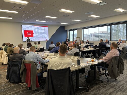 The image depicts a professional meeting or conference taking place in a modern, well-lit conference room. The room is filled with numerous individuals seated around several rectangular tables arranged in a U-shape. The attendees are predominantly dressed in business attire, indicating a formal or corporate setting. 

At the front of the room, a large screen displays a presentation slide with the title "Current Reality" in bold red text, suggesting a discussion or lecture on current issues or strategies. Two presenters stand near the screen, engaging with the audience. The tables are equipped with nameplates, notepads, and beverages, indicating an organized and structured event.

The room features large windows on one side, allowing natural light to illuminate the space and offering a view of trees and a distant building, adding an open and airy feel to the environment. The ceiling is fitted with recessed lighting, contributing to the bright and professional atmosphere.

The overall mood of the image is focused and collaborative, with participants appearing attentive and engaged in the proceedings. The setting suggests a corporate workshop, seminar, or strategic planning session, emphasizing themes of learning, collaboration, and professional development.