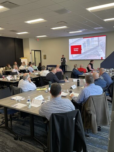 The image depicts a professional meeting or seminar taking place in a modern conference room. The setting includes several tables arranged in a U-shape, with participants seated around them. Each participant has a nameplate, suggesting a formal or organized event. The attendees are mostly dressed in business attire, indicating a corporate or business environment.

A speaker stands at the front of the room, presenting information displayed on a large screen. The screen shows a slide with the title "Current Reality," accompanied by text and a red rectangular graphic. This suggests the presentation might be addressing current business challenges or strategies.

The room is well-lit with fluorescent lighting, and the walls are painted in neutral tones, contributing to a professional atmosphere. Bottles of water and notepads are placed on the tables, indicating that the participants are engaged in note-taking or discussion.

The overall atmosphere is focused and attentive, with participants appearing engaged in the presentation. The setting and context suggest a business meeting, workshop, or training session aimed at discussing strategies, updates, or educational content relevant to the attendees' professional interests.
