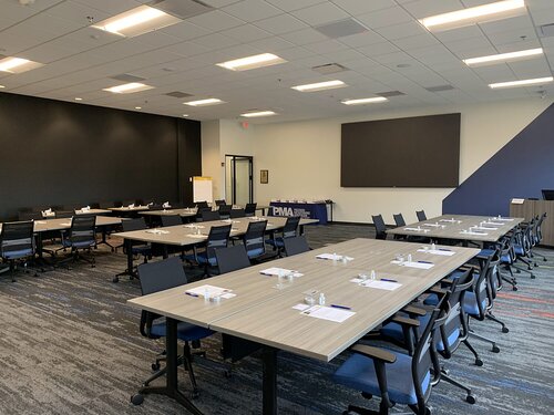 The image depicts a modern conference room or training facility with a professional and organized atmosphere. The room is set up with several rectangular tables arranged in a U-shape, surrounded by ergonomic office chairs. Each table is equipped with writing materials, including notepads and pens, suggesting a setting prepared for a meeting, seminar, or workshop.

The room features a neutral color palette, with shades of gray and blue dominating the decor. The flooring is carpeted with a subtle pattern that incorporates these colors, adding a touch of sophistication. The walls are painted in a light color, enhancing the room's brightness and spaciousness, while one wall is partially covered with a large blackboard or presentation screen, indicating a space designed for visual presentations or lectures.

Ceiling lights are evenly distributed, providing ample illumination throughout the room. At the back, a table is covered with a blue cloth, possibly used for refreshments or additional materials. The presence of a flip chart stand near the wall further emphasizes the room's purpose for interactive and educational activities.

Overall, the setting conveys a sense of professionalism and readiness for a corporate or educational event, with a focus on functionality and comfort.