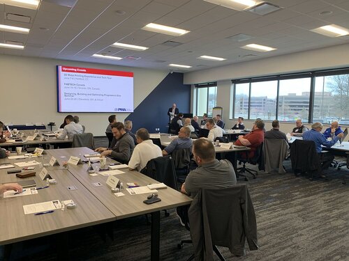 The image depicts a professional conference or meeting setting within a modern conference room. The room is filled with numerous attendees seated at rectangular tables arranged in a U-shape, allowing for interaction and discussion. Each table has nameplates and papers, indicating an organized and formal event. 

At the front of the room, a large screen displays a presentation slide titled "Upcoming Events," with a list of events including "FASTER," "Spring Meeting Experience and Plant Tour," and "Designing, Building, and Optimizing Progressive Dies." The screen is prominently positioned for all attendees to view. A speaker stands near the screen, likely presenting or addressing the group.

The room is well-lit with natural light streaming in from large windows on the right side, providing a view of an urban landscape with buildings and trees. The interior lighting is bright, enhancing the professional atmosphere. The carpet is dark with a subtle pattern, and the walls are neutral, contributing to a modern and business-like environment.

The attendees, predominantly dressed in business casual attire, are engaged, either listening to the speaker or discussing among themselves. The overall atmosphere is focused and collaborative, indicative of a professional development or industry-related event.

The setting and context suggest a corporate or industry conference, possibly related to manufacturing or engineering, given the topics on the presentation slide. The event is likely aimed at networking, knowledge sharing, and professional growth within a specific industry sector.