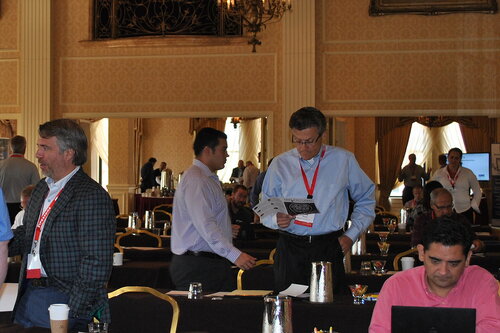 The image depicts a conference or seminar setting within an elegant, possibly historic venue. The room features ornate decor, including large chandeliers, tall columns, and gold accents, suggesting a formal or upscale environment. The lighting is warm, enhancing the luxurious ambiance.

Several individuals are engaged in various activities typical of a professional gathering. One man in the foreground is seated at a table, focused on a laptop, indicating active participation or note-taking. Another man stands nearby, examining a piece of paper, possibly a program or agenda, suggesting he is reviewing information pertinent to the event. A few other attendees are scattered throughout the room, either standing or seated, some conversing with each other.

The attendees are dressed in business or business-casual attire, with many wearing name tags on red lanyards, which is common at conferences for identification and networking purposes. Tables are set with water pitchers and glasses, emphasizing the provision of refreshments for participants.

The atmosphere is professional and collaborative, with a focus on learning or networking. The presence of screens and presentation equipment in the background hints at presentations or discussions taking place. The setting and activities suggest a business conference, workshop, or seminar, where attendees are likely exchanging ideas and gaining insights related to their field.