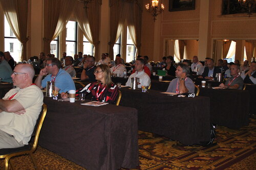 The image depicts a conference or seminar setting within an elegant, well-lit room. The room features large windows with cream-colored drapes, allowing natural light to illuminate the space. The walls are adorned with classic decor, including chandeliers and framed artwork, contributing to a sophisticated atmosphere.

The attendees, a diverse group of adults, are seated at long tables covered with dark tablecloths. They appear engaged, listening attentively to a presentation or speaker not visible in the image. Many individuals have notepads, pens, and drinks, suggesting a formal and educational environment. Some attendees wear lanyards, indicating they are part of an organized event, possibly a professional conference or workshop.

The predominant colors in the image include the warm tones of the carpet and walls, contrasted by the darker tablecloths and clothing of the attendees. The setting conveys a professional and focused atmosphere, typical of business or academic gatherings.