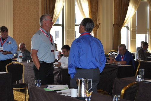 The image depicts a professional conference or meeting setting, likely taking place in a hotel or conference room. The room is well-lit with natural light streaming in through large windows, which are partially covered by elegant drapes. The walls are adorned with classic wallpaper, contributing to a formal and sophisticated atmosphere.

In the foreground, two men are engaged in conversation. One is wearing a light gray polo shirt, while the other is in a bright blue dress shirt. Both have conference badges hanging around their necks, indicating they are participants or speakers at the event. Their body language suggests a friendly and engaged discussion.

The room is filled with several tables covered in dark tablecloths, each equipped with water glasses, pitchers, and notepads, suggesting a setup for note-taking or presentations. Other attendees are seated at these tables, some looking at their phones or writing notes, indicating a break or informal networking period.

The predominant colors in the image are warm tones from the walls and curtains, contrasted by the dark tablecloths and the bright clothing of the attendees. The overall atmosphere is professional and collegial, typical of a business or industry conference.

The image captures a moment of networking and exchange of ideas, common in professional gatherings, without any specific cultural or entertainment references.
