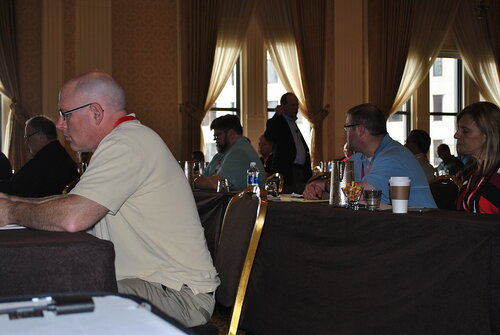 The image depicts a conference or seminar setting with several individuals seated at tables, facing forward, suggesting they are listening to a speaker or presentation. The room is elegantly decorated, featuring tall windows with sheer curtains that allow natural light to filter in, creating a warm and inviting atmosphere. The lighting is soft, primarily from the natural light, complemented by the room's ambient lighting.

The attendees are dressed in business casual attire, indicating a professional yet relaxed environment. On the tables, there are various items such as notepads, pens, water bottles, and coffee cups, suggesting the participants are engaged in note-taking or discussion. A few individuals have lanyards around their necks, which are typical at conferences for identification purposes.

The predominant colors in the image are neutral tones, with beige and brown hues from the walls and furniture, contrasted by the colorful attire of the attendees. The overall mood is focused and attentive, typical of a learning or networking event.

In the background, a person is standing, possibly an organizer or speaker, adding to the dynamic of the scene as they may be addressing the audience or preparing to do so. The setting suggests a formal gathering, likely related to business, education, or professional development.