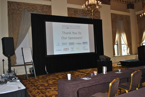 The image depicts a conference or seminar setting, likely in a hotel ballroom or similar venue. The room features ornate decor, including chandeliers and heavy drapery, suggesting a formal or upscale environment. The predominant colors are warm tones, with gold and brown hues in the carpet and chairs.

At the center is a large projection screen displaying a slide that reads "Thank You To Our Sponsors!" followed by a list of sponsor logos. These logos include recognizable brands such as Infor, IQMS, and HubSpot, among others. This indicates the event is likely related to business, manufacturing, or marketing.

In front of the screen, there are several rows of tables and chairs arranged classroom-style, suggesting a presentation or lecture format. On the tables are items like water pitchers and glasses, typical of conference setups. The lighting is bright, focused on the screen and stage area, enhancing visibility for attendees.

The atmosphere is professional and business-oriented, with an emphasis on networking and information sharing. The presence of technical equipment, such as a projector and audio-visual setup, further supports the context of a formal presentation or workshop.