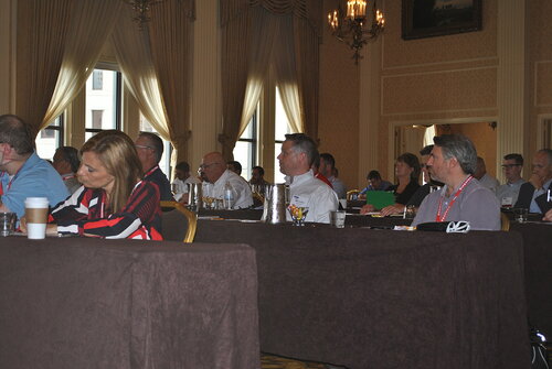 The image depicts a conference or seminar setting in a formal, well-lit room with elegant decor. The room features high ceilings, ornate chandeliers, and large windows with draped curtains, suggesting a venue of historical or upscale significance. Attendees are seated at tables covered with dark brown tablecloths, arranged in rows facing a direction outside the frame, indicating a focus on a speaker or presentation.

The individuals in the image are predominantly dressed in business or business-casual attire, such as collared shirts and blouses, which aligns with a professional event. Some attendees have lanyards around their necks, likely indicating name tags or event passes. A woman in the foreground wears a red, black, and white striped top, adding a splash of color to the scene.

On the tables, there are various items such as water bottles, glasses, and notepads, suggesting that participants are engaged in note-taking or discussion. A few disposable coffee cups are visible, indicating refreshments are available.

The atmosphere is focused and attentive, with most individuals appearing to listen intently or take notes. The setting and attire suggest a formal business conference or workshop, potentially related to corporate training, industry networking, or a professional symposium. The image conveys a sense of professionalism and engagement within a structured learning or networking environment.