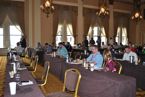 The image depicts a conference or seminar setting within an elegant, historic-style room. The room features ornate architectural details, including high ceilings, large windows with sheer curtains, and intricate chandeliers that provide warm lighting. The walls are adorned with patterned wallpaper, contributing to a classic and sophisticated atmosphere.

Attendees are seated at long tables covered with brown tablecloths, arranged in rows facing the front of the room. The tables are equipped with notepads, pens, water bottles, and coffee cups, indicating a formal meeting or presentation is taking place. The participants, a mix of men and women, are casually dressed, suggesting a business-casual or informal dress code. Some individuals are engaged in conversation, while others appear to be listening attentively or taking notes.

The overall color palette of the room includes warm tones of gold, brown, and beige, which complement the natural light streaming in from the windows. The atmosphere is professional yet relaxed, typical of a corporate or educational event.

The setting and decor suggest a venue that could be part of a historic hotel or conference center, often used for business meetings, workshops, or training sessions. The presence of modern technology, such as laptops and projectors, indicates a blend of traditional and contemporary elements, suitable for a variety of professional gatherings.