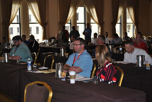 The image depicts a conference or seminar setting in a large, well-lit room with multiple rows of tables and chairs. The room features tall windows with curtains, allowing natural light to illuminate the space. Attendees, both men and women, are seated at the tables, which are covered with dark tablecloths. They appear to be listening attentively, taking notes, or engaging in quiet conversation. 

Several individuals are wearing name tags, suggesting a formal or professional gathering. On the tables, there are various items such as notebooks, pens, water bottles, and a Starbucks coffee cup, indicating a focus on note-taking and refreshment. A few glass pitchers and bowls of candy are also visible, providing additional amenities for the attendees.

The attire of the participants is predominantly business casual, with some wearing collared shirts and others in more casual tops. The atmosphere is professional and focused, typical of a corporate or educational event. The room's decor, with its elegant curtains and gold-framed chairs, adds a touch of formality to the setting.

Overall, the image conveys a sense of a structured and organized event, likely aimed at professional development, networking, or knowledge sharing.
