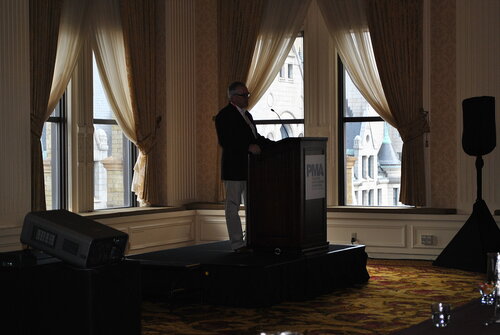 The image depicts a formal indoor setting, likely a conference or seminar, with a man standing at a podium. The room features large, elegant windows with sheer curtains, allowing natural light to filter in, casting a soft glow. The walls are adorned with ornate patterns, and the carpet has a rich, intricate design, contributing to a sophisticated atmosphere.

The man at the podium is dressed in formal attire, including a dark jacket and light-colored pants, suggesting a professional or business event. The podium has a sign with the acronym "PWNA," indicating the event might be related to an organization or association with those initials.

The setting is a large room with high ceilings, enhancing the grandeur of the space. The lighting is subdued, with the focus on the speaker, creating an intimate yet formal ambiance. The presence of a projector suggests the possibility of a presentation or lecture being part of the event.

Overall, the image conveys a sense of professionalism and formality, typical of corporate gatherings or industry conferences. The architectural details and decor reflect a classic and upscale environment, suitable for significant meetings or presentations.