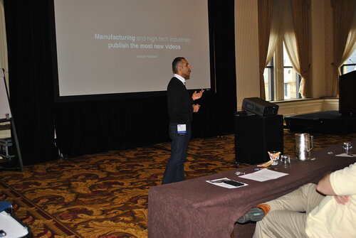 The image depicts a conference or seminar setting in a well-lit room with ornate decor, including patterned carpet and large windows with drapes. A man is standing in front of a large projection screen, giving a presentation. He is dressed in a black sweater and jeans, with a visible name badge clipped to his waistband. The screen displays a slide with the text: "Manufacturing and high tech industries publish the most new videos" and attributes the quote to "Shout Marketing."

In the foreground, there is a table with a notepad, pen, and a glass of water, suggesting a formal or professional environment. The audience, partially visible, is seated and appears to be attentive, with one person leaning forward, possibly taking notes or engaging with the presentation.

The atmosphere is professional and focused, indicative of a business or industry-related event. The setting suggests a workshop or conference room, possibly in a hotel or convention center, given the elegant interior design. The image conveys a theme of learning and information exchange, typical of industry conferences or seminars.