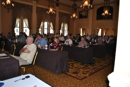The image depicts a conference or seminar setting in an ornate, classical-style room. The audience is seated at long tables covered with dark brown tablecloths, facing the front of the room. The attendees are predominantly adults, dressed in casual to business-casual attire, indicating a professional yet relaxed atmosphere. 

The room is elegantly decorated, featuring high ceilings with intricate moldings and large chandeliers that provide warm lighting. Tall windows with draped curtains allow natural light to complement the interior illumination. The walls are adorned with framed artwork, adding to the sophisticated ambiance.

The carpet is patterned with rich red and gold hues, enhancing the room's luxurious feel. Notebooks, pens, and water bottles are visible on the tables, suggesting that the participants are engaged in a learning or discussion session. The overall mood is focused and attentive, typical of a formal gathering aimed at knowledge sharing or professional development.

This setting is reminiscent of venues used for corporate events, academic conferences, or industry workshops, where networking and information exchange are key components. The combination of elegant decor and professional attire underscores the event's formal and educational nature.