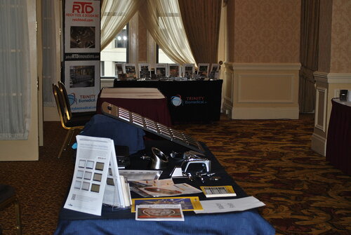 The image depicts an indoor conference or exhibition setting, characterized by a series of tables displaying various promotional materials and equipment. The room features patterned carpet flooring in shades of red and gold, and the walls are adorned with elegant wallpaper, contributing to a formal and professional atmosphere. 

In the foreground, a table is covered with a dark blue tablecloth, showcasing brochures, pamphlets, and what appears to be electronic equipment or medical devices. Prominent among the materials is a brochure stand displaying multiple flyers, suggesting a focus on informational distribution. 

In the background, another table is visible, draped with a black tablecloth branded with the logo and name "Trinity Biomedical." Behind this table, a series of framed photographs or certificates are arranged, possibly highlighting achievements or product offerings. 

Adjacent to this, a vertical banner displays the logo and website of "RTD," indicating a potential partnership or shared event space between the two entities. The banner and table setup suggest a trade show or professional networking event focused on biomedical or technological innovations.

Natural light filters through large, draped windows, adding a soft illumination to the scene. The overall mood is professional and focused, likely aimed at engaging attendees with the products and services on display.