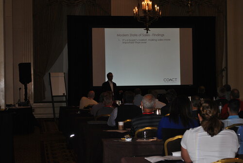 The image depicts a conference or seminar setting in a dimly lit room, where a presentation is taking place. The focal point is a large projection screen displaying a slide titled "Modern State of Sales - Findings." The slide lists a point: "1. It's a buyer's market; making sales more important than ever." The bottom right of the slide features the logo "COACT" with the tagline "Sales Agency."

In front of the screen, a speaker stands, silhouetted against the bright projection, addressing an audience seated at tables. The audience consists of men and women, some with visible notepads and pens, suggesting they are taking notes or engaged in the presentation. The tables are covered with dark tablecloths, and some attendees are wearing lanyards, indicating this is a formal event, possibly a business conference or workshop.

The room is decorated with elegant elements, such as chandeliers and draped curtains, suggesting a hotel or conference center setting. The lighting is subdued, focusing attention on the presentation screen. The atmosphere is professional and focused, with attendees appearing attentive and engaged in the content being delivered.

Overall, the image conveys a business-oriented theme, emphasizing the importance of sales strategies in a competitive market environment.