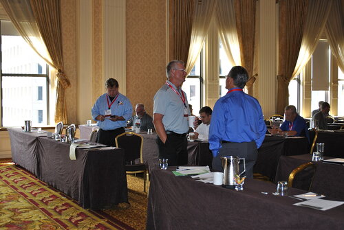 The image depicts a conference or meeting setting in a large, elegant room with high ceilings and ornate decor. The room features tall windows with sheer curtains, allowing natural light to illuminate the space. The walls are adorned with intricate patterns, contributing to a sophisticated atmosphere.

Several individuals are engaged in conversation or focused on their tasks. They are dressed in business or casual business attire, with some wearing name tags, indicating a formal event or gathering. The attendees are spread across tables covered with dark tablecloths, which are equipped with water pitchers, glasses, and papers, suggesting a seminar or workshop environment.

The carpet has a rich, patterned design in red and gold tones, complementing the overall opulent aesthetic of the room. The lighting is bright, primarily due to the natural light from the windows, creating a warm and inviting ambiance.

The scene conveys a professional and collaborative atmosphere, typical of business conferences or industry meetings. The individuals appear engaged and focused, contributing to a sense of productivity and networking.