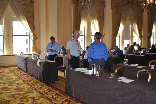 The image depicts a conference or seminar setting in an elegant, well-lit room with high ceilings and large windows. The room features ornate chandeliers and decorative curtains, contributing to a formal and sophisticated atmosphere. The carpet has a rich, intricate pattern with shades of red and gold, complementing the room's luxurious ambiance.

Several individuals are engaged in conversation, standing between rows of tables covered with dark brown tablecloths. Each table is equipped with notepads, papers, and water glasses, indicating a professional or educational event. The attendees are dressed in business attire, suggesting a corporate or industry-related gathering.

The lighting is bright, with natural light streaming in through the windows, enhancing the room's grandeur. The overall mood is professional and focused, with participants seemingly preparing for or discussing a presentation or workshop.

This setting is typical for business conferences, training sessions, or industry meetings, where networking and information exchange are key activities. The room's decor and setup reflect a formal and organized event, emphasizing professionalism and collaboration.