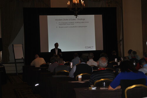 The image depicts a conference or seminar setting in a dimly lit room. A presenter stands at the front, silhouetted against a projected presentation slide. The slide is titled "Modern State of Sales - Findings" and lists two key points: "1. It’s a buyer’s market; making sales more important than ever" and "2. Buyers want consultative salespeople." The slide includes the logo "COACT" at the bottom right.

The audience consists of several individuals seated at tables, facing the presenter. They appear to be engaged, possibly taking notes or listening attentively. The room is decorated with chandeliers, adding a formal and professional atmosphere. The lighting is subdued, focusing attention on the presentation screen.

The overall mood is serious and business-like, indicative of a professional development or sales training event. The setting suggests a focus on learning and exchanging ideas related to sales strategies and market dynamics.