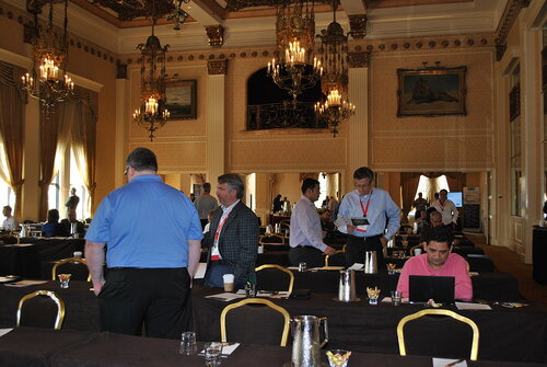 The image depicts a formal conference or seminar setting within an opulent, historic ballroom. The room features high ceilings adorned with ornate chandeliers, and the walls are decorated with classical paintings and intricate moldings, adding to the grandeur of the space. The lighting is warm and elegant, casting a sophisticated ambiance throughout the room.

Several individuals are scattered across the room, engaging in conversation or working on laptops, suggesting a break or networking session during the event. The attendees are dressed in business casual attire, indicating a professional gathering. Tables are arranged in rows, covered with dark tablecloths, and equipped with notepads, pens, and water pitchers, typical of a conference setup.

The atmosphere is one of focused engagement and professional interaction, with a mix of people standing and seated, contributing to a dynamic yet orderly environment. The presence of name tags on some individuals suggests a formal registration process for the event.

Overall, the image conveys a sense of a high-profile business or academic conference taking place in a luxurious, historic venue, emphasizing networking and professional development.