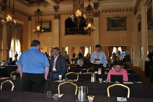 The image depicts a conference or seminar setting in an opulent, chandelier-lit room with ornate decorations and high ceilings. The room features elegant, classical architecture with large windows draped in heavy curtains, adding to the grandeur. The walls are adorned with framed artwork, and the lighting is warm, creating a sophisticated atmosphere.

Several individuals are engaged in conversation or working at tables covered with dark tablecloths. One person in a blue shirt is standing and conversing with another in a checkered blazer, suggesting a professional or networking interaction. Another individual is seated, focused on a laptop, indicating a work-related activity. Attendees are wearing name tags on red lanyards, typical of conference events.

The tables are equipped with conference essentials such as water pitchers, cups, and small bowls of snacks, indicating a formal meeting or presentation environment. The presence of a projector screen in the background suggests that presentations or speeches might be part of the event.

The overall atmosphere is professional and collaborative, with an emphasis on networking and information exchange, typical of business or academic conferences. The setting and attire of the attendees convey a formal and serious tone, with a focus on engagement and learning.