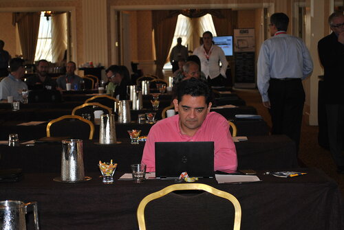 The image depicts a conference or seminar setting in a well-lit room with several rows of tables and chairs. The room has an elegant decor, featuring gold-framed chairs and dark tablecloths. In the foreground, a man in a pink shirt is seated at a table, focused on his laptop. The table is equipped with a glass bowl of colorful candies, a notepad, and a pen, suggesting a formal or educational event.

In the background, several other attendees are engaged in conversations or working on their laptops. Some individuals are wearing name tags, indicating a professional gathering. The lighting is warm, with natural light filtering through large windows adorned with drapes, creating a welcoming atmosphere.

Prominent objects include metallic water pitchers and glasses on the tables, reinforcing the formal nature of the event. A display board with text is visible in the background, likely providing information about the event or sponsors.

The overall mood is professional and focused, with participants engaged in activities typical of a conference or workshop. The setting and attire suggest a business or academic environment, with a mix of networking and learning activities taking place.
