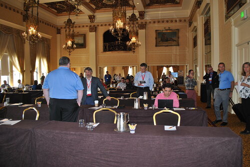 The image depicts a conference or seminar setting in an opulent, historic-style ballroom. The room features high ceilings adorned with ornate chandeliers and intricate moldings. Large windows with elegant drapery allow natural light to illuminate the space, complementing the warm, ambient lighting from the chandeliers. The walls are decorated with framed paintings, adding to the sophisticated atmosphere.

In the foreground, several rows of tables are covered with dark tablecloths. On the tables are items typical of a conference setting, such as glasses, pitchers, and small bowls of candy. Attendees, predominantly dressed in business casual attire, are engaged in various activities. Some are standing and conversing, while others are seated, focused on their laptops or notes. A few individuals wear lanyards with name tags, indicating their participation in the event.

The room is bustling with activity, suggesting a break or networking session. The overall atmosphere is professional yet relaxed, with a sense of collaboration and exchange of ideas. The presence of technology, such as laptops and projectors, hints at a modern conference theme, possibly related to business, technology, or education.

The setting, with its blend of historical architecture and contemporary conference elements, creates a unique juxtaposition, highlighting the fusion of tradition and modernity.