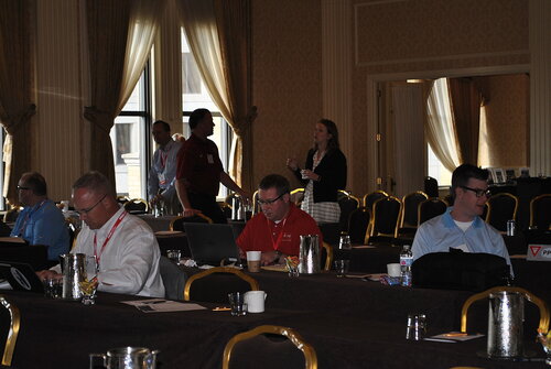 The image depicts a conference or seminar setting within a formal, well-lit room. The room features ornate decor with large windows draped with curtains, allowing natural light to filter in. The atmosphere is professional and focused, suggesting a business or educational event.

Several individuals are seated at tables covered with dark tablecloths, working on laptops or engaging in conversation. The tables are equipped with water bottles, coffee cups, and notepads, indicating a setting designed for note-taking and discussions. Some attendees are wearing lanyards, which are typical for conferences, suggesting they are participants or speakers.

In the background, a group of people is standing and conversing, indicating networking or a break between sessions. The individuals are dressed in business casual attire, which is common for such events. The room is filled with rows of chairs, hinting at a larger audience capacity.

The lighting is a mix of natural and artificial, creating a warm and inviting ambiance. The overall mood is one of concentration and engagement, typical of a professional gathering aimed at learning or collaboration.