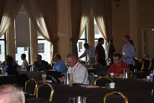 The image depicts a conference or seminar setting in a large, elegant room with high ceilings and tall windows draped with sheer curtains, allowing natural light to filter in. The room is furnished with rows of tables covered in dark tablecloths, and each table is equipped with water pitchers, glasses, and various personal items like laptops and notebooks.

Several individuals are seated at the tables, engaged in activities such as typing on laptops, writing notes, or conversing with one another. A few people are standing, possibly networking or discussing topics related to the event. Most attendees are wearing business casual attire, and many have lanyards with name tags, indicating their participation in the event.

The atmosphere is professional and focused, with a sense of collaboration and learning. The lighting is a mix of natural daylight from the windows and warm indoor lighting, creating a comfortable environment. The setting suggests a formal gathering, such as a business conference, workshop, or training session.

The room's decor, including ornate wallpaper and gold-framed chairs, adds a touch of sophistication and formality to the event. The presence of technology, such as laptops and smartphones, indicates a modern and connected atmosphere, likely involving discussions or presentations on contemporary topics.