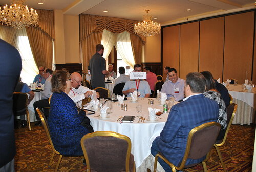 The image depicts a formal dining setting, likely a conference or business meeting, held in an elegant banquet room. The room features ornate chandeliers and large windows with draped curtains, contributing to a sophisticated atmosphere. Several round tables are set with white tablecloths, neatly arranged cutlery, glassware, and folded napkins, indicating a meal is either ongoing or about to commence.

At one of the tables, a group of individuals is engaged in conversation. They are dressed in business attire, suggesting a professional event. A small sign on one of the tables reads "Educational Track," indicating that this might be a breakout session or themed discussion within a larger conference.

The predominant colors in the room are warm tones, with gold and beige hues in the carpet and curtains, complemented by the white table settings. The lighting is bright, provided by both the chandeliers and natural light from the windows, creating an inviting and formal ambiance.

The atmosphere is professional and collegial, with attendees appearing to be networking or discussing business-related topics. The setting and attire suggest this is a corporate or educational event, possibly related to a seminar, workshop, or industry conference.