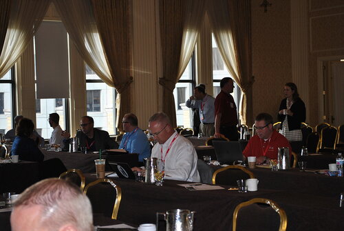The image depicts a conference or seminar setting within a well-lit room, characterized by large windows with sheer curtains allowing natural light to filter in. The room features ornate decor, with patterned wallpaper and gold-accented chairs, suggesting a formal or upscale venue.

Several individuals are seated at long tables covered with dark tablecloths, engaged in activities such as typing on laptops, writing notes, or conversing. The attendees are dressed in business casual attire, with some wearing lanyards, indicating a professional event. Coffee cups, water bottles, and notepads are scattered across the tables, suggesting a working session or presentation.

The atmosphere is focused and attentive, with participants appearing engaged in the proceedings. The lighting is a mix of natural daylight from the windows and interior lighting, creating a balanced and inviting environment. The setting and activities suggest a corporate meeting, workshop, or training session, possibly related to business or technology.

Overall, the image conveys a professional and collaborative atmosphere, with a clear emphasis on learning and interaction among the attendees.