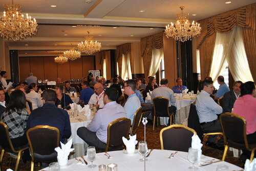 The image depicts a formal dining event taking place in an elegant banquet hall. The room is adorned with ornate chandeliers that provide warm, ambient lighting, enhancing the sophisticated atmosphere. The walls are lined with large windows draped with luxurious, gold-colored curtains, allowing natural light to complement the interior lighting.

The attendees, a diverse group of individuals, are seated at round tables covered with white tablecloths. Each table is set with neatly arranged silverware, glasses, and napkins, indicating a formal dining setup. The participants appear engaged in conversation, suggesting a social or networking event, possibly a conference or corporate gathering.

The overall color palette of the room includes neutral tones with accents of gold and white, contributing to the upscale ambiance. The attire of the individuals ranges from business casual to formal, aligning with the professional setting.

The setting and arrangement suggest a structured event, likely involving presentations or discussions, as indicated by the presence of a projector screen and audio equipment visible in the background. The image conveys a sense of professionalism and camaraderie, typical of corporate or formal social events.
