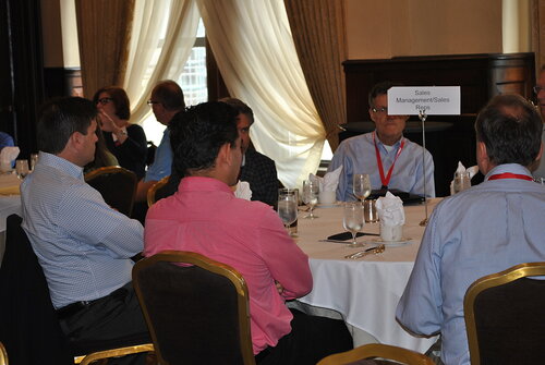 The image depicts a professional conference or meeting setting, likely a business event, as suggested by the formal attire of the attendees and the organized table setup. Several individuals are seated around a round table, engaged in conversation or listening attentively. The table is covered with a white tablecloth and set with glasses, napkins, and cutlery, indicating a formal dining arrangement.

A prominent sign on the table reads "Sales Management/Sales Reps," suggesting that the discussion may be focused on sales strategies or networking among sales professionals. The participants are wearing business casual attire, with some in dress shirts and others in blazers, and a few have name tags or lanyards, which is typical for conferences.

The room features elegant decor, with large windows draped with sheer curtains that allow natural light to filter in, creating a warm and inviting atmosphere. The lighting is soft, contributing to a professional yet relaxed ambiance. The chairs are upholstered in a dark fabric, complementing the room's classic and sophisticated style.

The setting suggests a collaborative and focused environment, where individuals are likely sharing insights, strategies, or networking. The presence of multiple tables and attendees indicates a larger event, possibly a workshop or seminar, aimed at professional development within the sales industry.