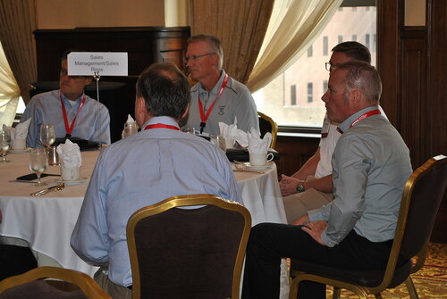 The image depicts a professional setting, likely a conference or business meeting, taking place in a well-appointed room with large windows and elegant drapery. A group of five men are seated around a round table, engaged in what appears to be a discussion or presentation. Each individual is wearing a name badge on a red lanyard, suggesting an organized event where identification is necessary.

The table is covered with a white tablecloth and set with glasses, napkins, and silverware, indicating a formal dining setup, possibly for a luncheon or dinner meeting. A sign on the table reads "Sales Management/Sales Reps," designating the group or topic of discussion. The men are dressed in business casual attire, with collared shirts and no jackets, appropriate for a professional yet relaxed environment.

The room is warmly lit, contributing to a professional and inviting atmosphere. The presence of the sign and the attire of the individuals suggest a corporate or sales-related event, where networking and information exchange are key activities. The overall mood is one of focus and engagement, typical of a business meeting or seminar setting.