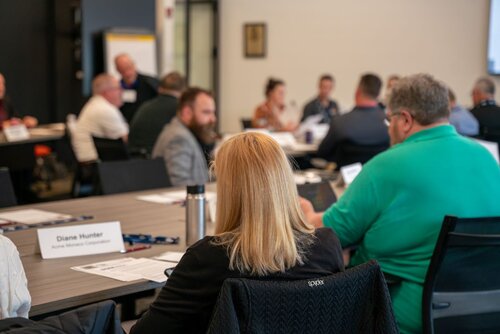 The image depicts a professional meeting or conference setting, likely a workshop or seminar. The room is filled with people seated around tables, engaged in discussion or listening to a speaker. The individuals are dressed in business casual attire, suggesting a formal yet relaxed atmosphere. 

Prominent objects include name tags and documents on the tables, indicating an organized event. One visible name tag reads "Diane Hunter" from "Acme Widget Company," suggesting a business context. A stainless steel water bottle is also noticeable on the table, hinting at a long session where attendees need refreshments.

The setting appears to be a conference room with neutral-colored walls and modern furnishings. The lighting is bright and even, typical of indoor professional environments. 

The atmosphere is focused and collaborative, with participants likely exchanging ideas or learning from a presentation. The image conveys a sense of professionalism and networking, common in corporate or educational events.