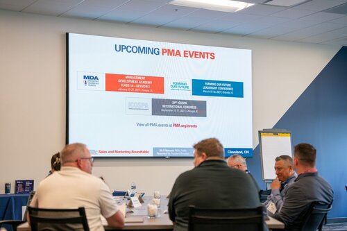 The image depicts a professional meeting or conference setting. In the foreground, several individuals are seated around a table, engaged in discussion. They are dressed in business casual attire, suggesting a formal yet relaxed atmosphere. The table is equipped with water bottles, notepads, and pens, indicating a focus on note-taking and active participation.

Prominently displayed on the wall is a large screen showing a presentation slide titled "UPCOMING PMA EVENTS." The slide lists three events: "Management Development Academy Class 5 - 25 Sessions," "ICOSPA 22nd International Congress," and "Forming Our Future Leadership Conference." Each event is associated with a different theme or focus, as indicated by the color-coded sections on the slide. The events appear to be related to professional development and industry leadership.

The room is well-lit with overhead fluorescent lighting, creating a bright and focused environment. The walls are painted in neutral tones, contributing to a professional setting. In the background, a flip chart is visible, further emphasizing the meeting's collaborative and strategic nature.

The location is identified as Cleveland, OH, as noted on the presentation slide. This detail, along with the specific events listed, suggests a context of industry networking and professional growth, likely within a business or manufacturing sector.

Overall, the image conveys a theme of professional development and industry collaboration, with a focus on upcoming events designed to enhance skills and leadership within the field.