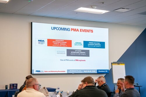 The image depicts a conference room setting where a group of individuals is engaged in a meeting or presentation. The focal point is a large screen displaying information about upcoming PMA (Precision Metalforming Association) events. The screen is divided into sections with details about different events:

1. **Management Development Academy**: Scheduled for January 24-25, 2024, in Nashville, TN, and March 6-7, 2024, in Cleveland, OH.

2. **Forming Our Future Leadership Conference**: Taking place from March 6-9, 2024, in Scottsdale, AZ.

3. **21st ICOSPA International Congress**: Scheduled for September 23-27, 2024, in Chicago, IL.

The bottom of the screen provides a link to view all PMA events at PMA.org/events.

The setting is a modern conference room with neutral tones, predominantly blue and gray, creating a professional atmosphere. Attendees are seated around tables, some taking notes, suggesting an educational or strategic planning context. A flip chart is visible, indicating a collaborative or interactive session.

The image conveys a business-like, organized environment focused on professional development and industry networking. The lighting is bright and even, typical of a corporate setting, enhancing the clarity of the presentation.
